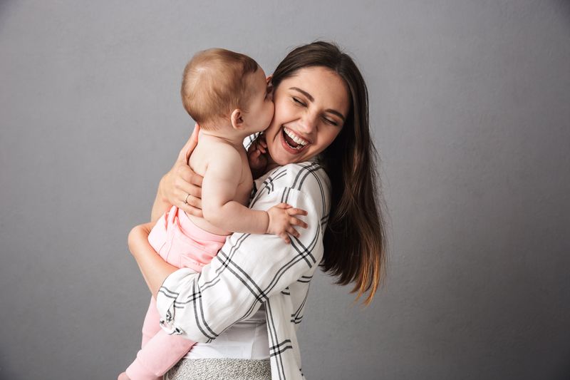 Portrait of a joyful young mother holding her little baby girl over gray background